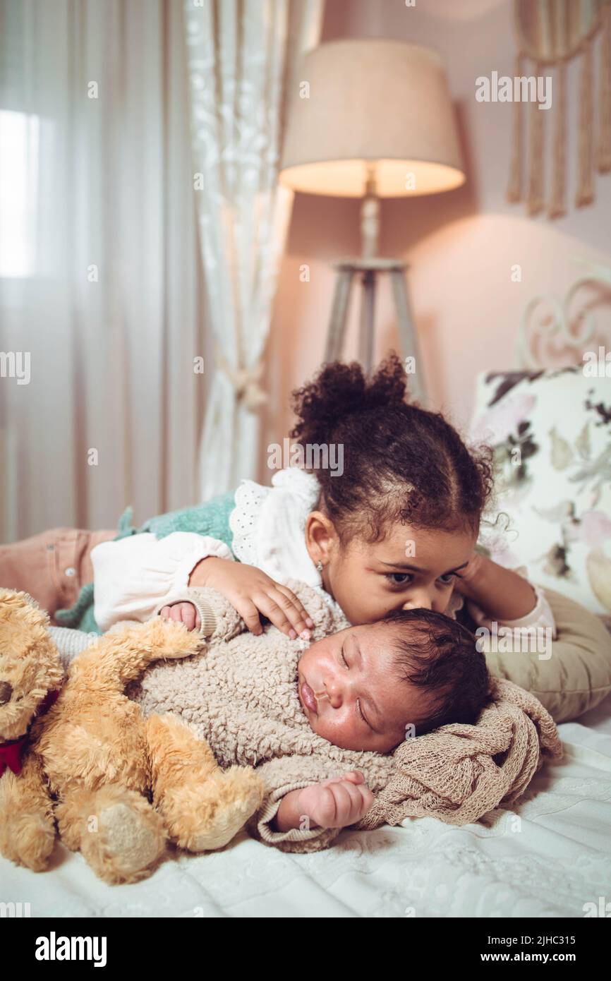 Little multiethnic girl, sister kissing her baby infant brother while they are lying on the bad Stock Photo