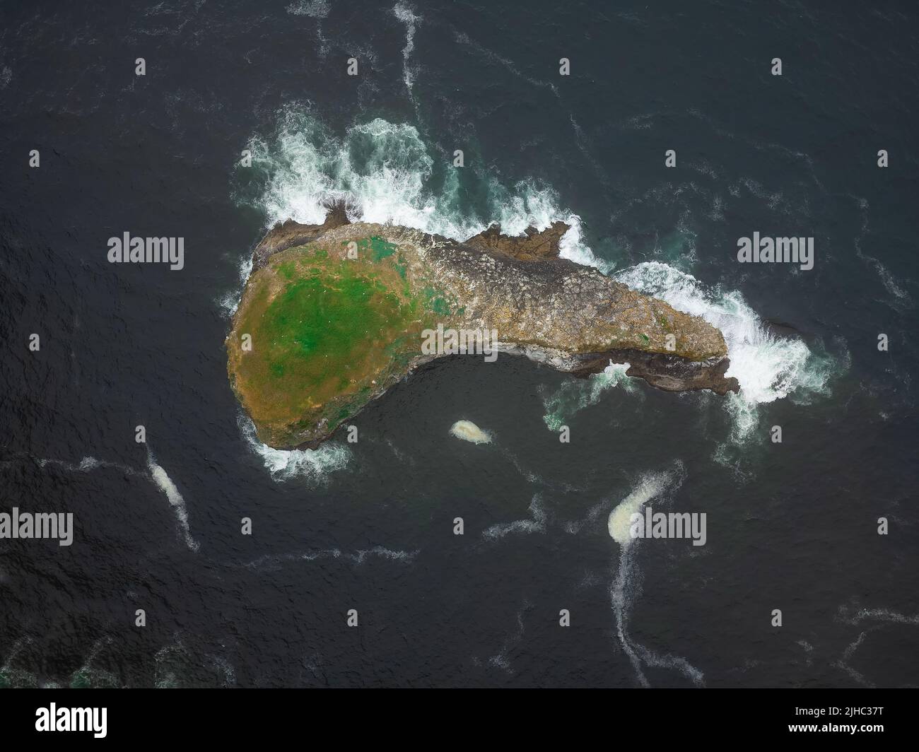 Shooting from a drone. A bizarre stone island in the ocean in the shape of a fish, covered with moss. Beauty of nature. There are no people in the pho Stock Photo