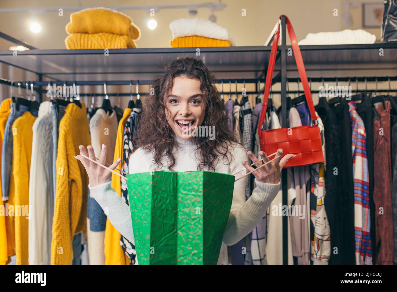 Shopaholic. Beautiful young girl woman in a clothing store in the mall rejoices shoppers gift. Stands and holds a green paper bag on a background of hangers with clothes. He looks at