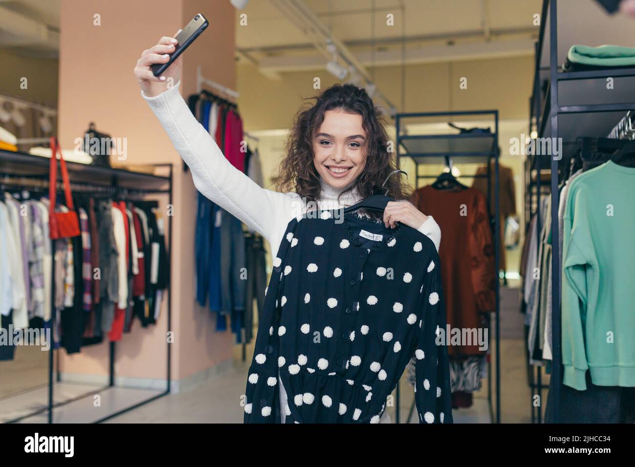 Young beautiful happy woman girl trying on a black dress with white dots near the mirror in a clothing store in the mall. Takes a selfie, shows friends, calls on a video call. He smiles, wants to buy Stock Photo