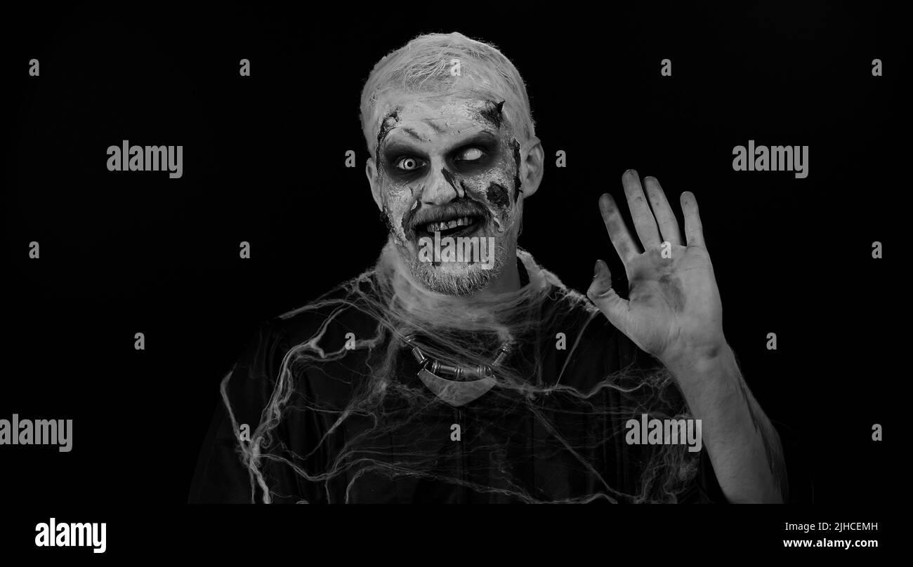 Creepy man with bloody scars face, Halloween zombie make-up. Scary wounded undead guy smiling friendly at camera and waving hands gesturing hello or goodbye, welcoming with hospitable expression Stock Photo