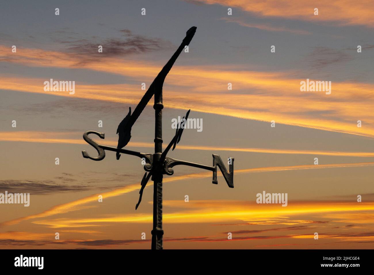 A weather vane points to the west with a sunset sky in the background in a composite image. Stock Photo