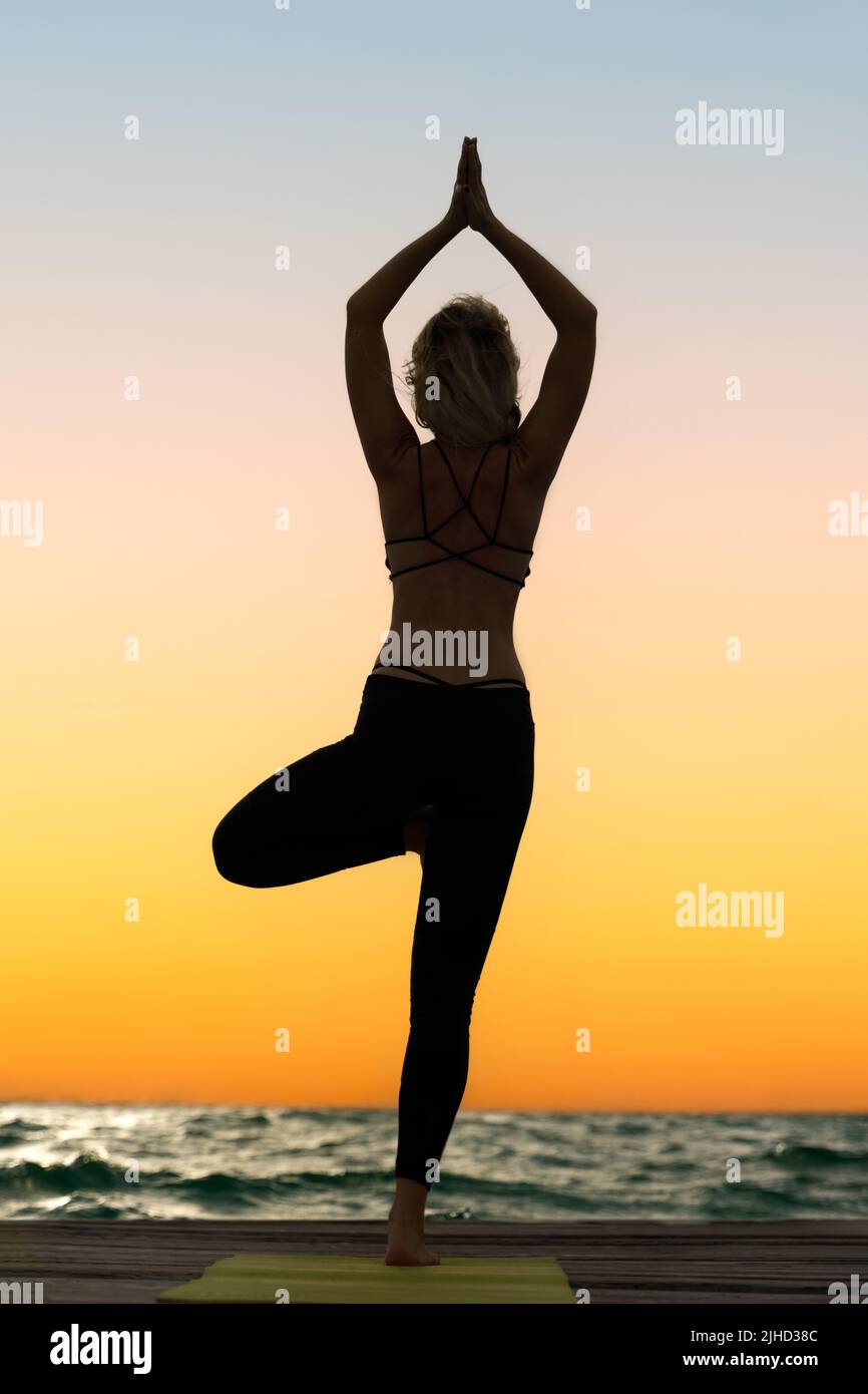 A young woman practices yoga on the ocean coast during sunrise. Stock Photo