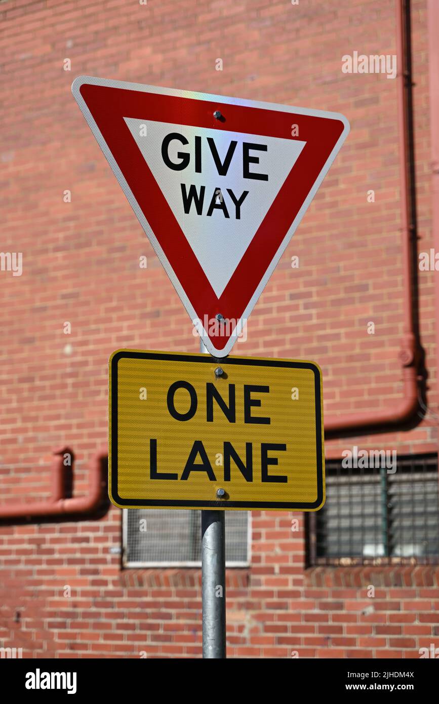 A red and white give way sign, featuring black text, above a yellow and black one lane sign, with a red brick building in the background Stock Photo