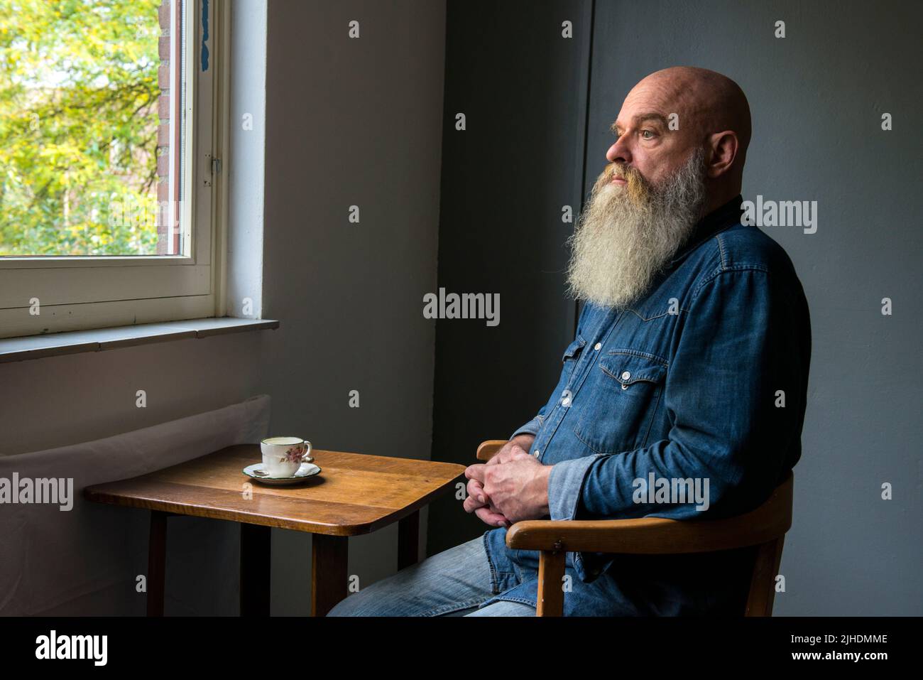 Tilburg, Netherlands. Portrait of a mature adult, caucasian male sitting next to a coffee table, drinking a cup of tea or coffee, meanwhile being in a zen state contemplating life and enjoying the view through the window. Stock Photo