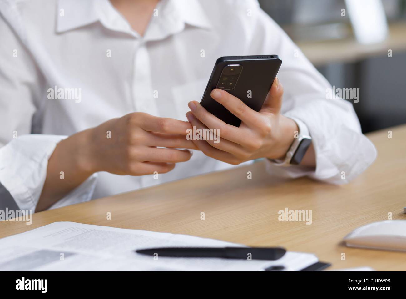 Close-up photo. The hands of a young woman in a white shirt are holding a mobile phone, typing a message at the table. Stock Photo