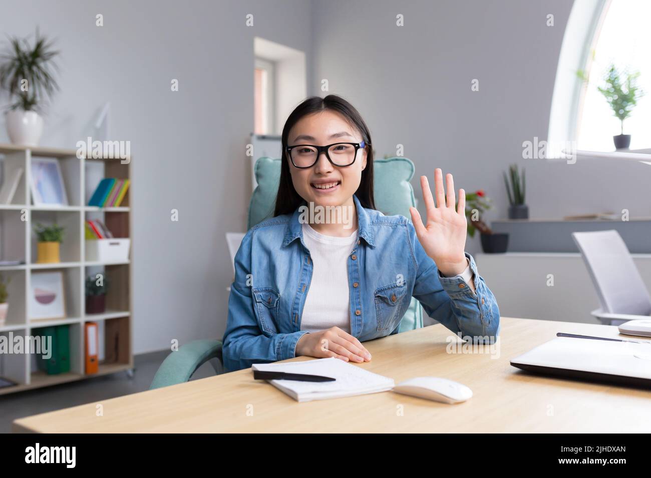 Distance Learning. A young Asian woman is a teacher, conducts online classes, lessons. He looks into the camera, explains, tells. Sitting in glasses and jeans at a table at a computer in a classroom Stock Photo