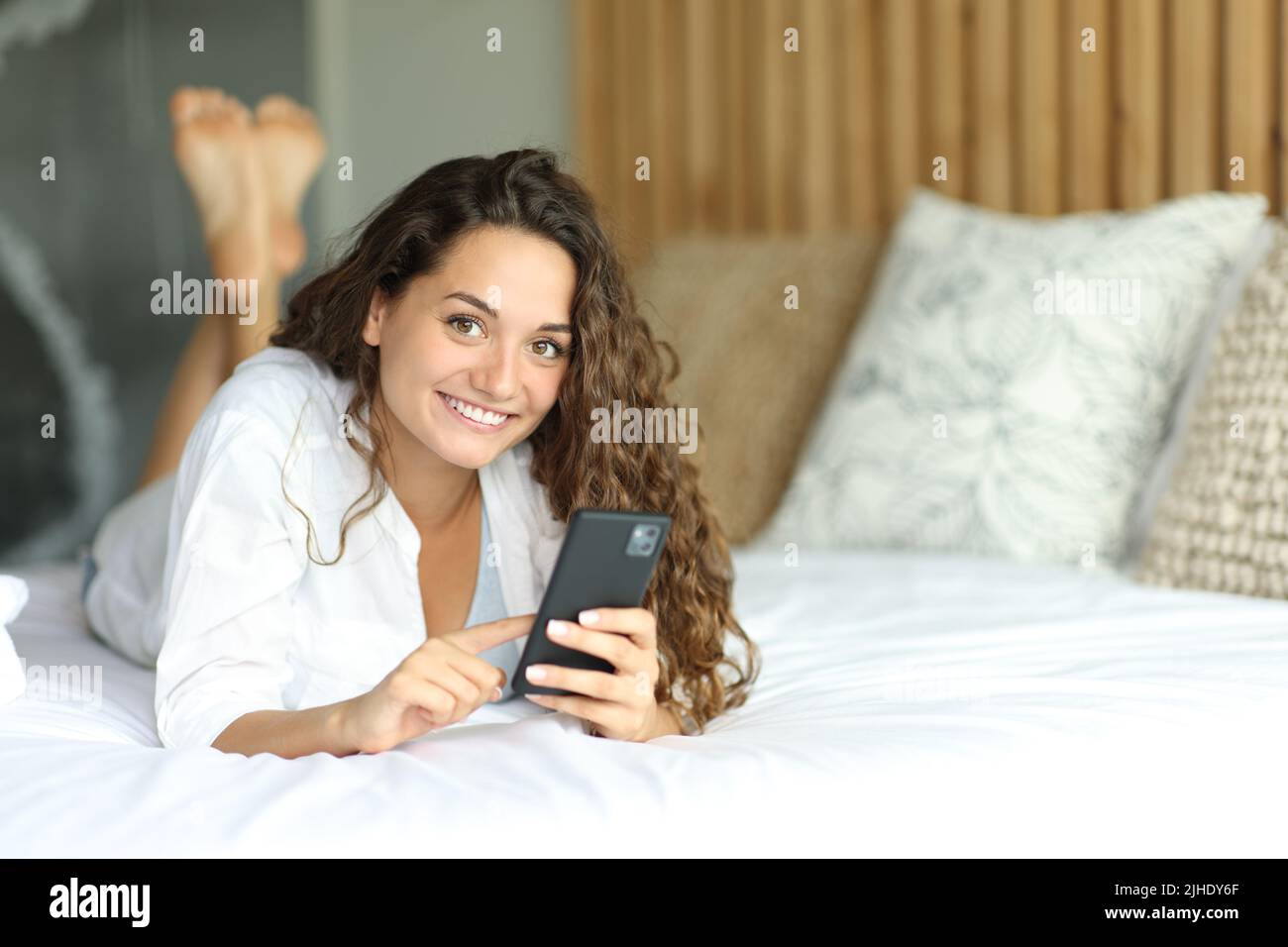 Happy woman holding smart phone looks at you lying on a bed Stock Photo