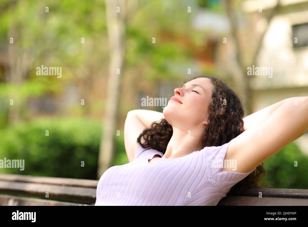 Relaxed woman resting with arms on head sitting in a bench in a park ...