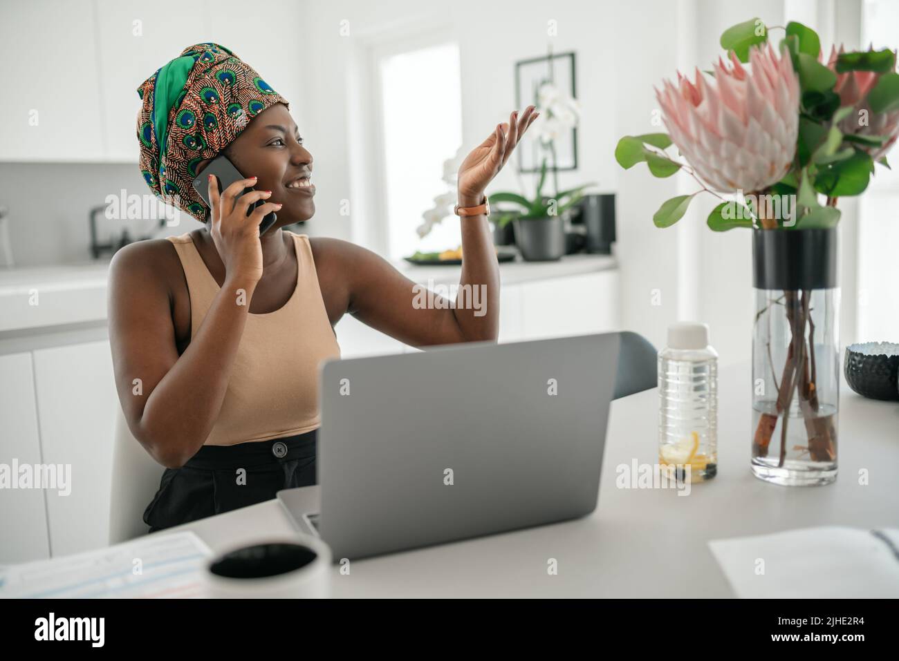 Beautiful young Black African woman wearing tradition headscarf. Sitting at home working on laptop. On phone call smiling Stock Photo