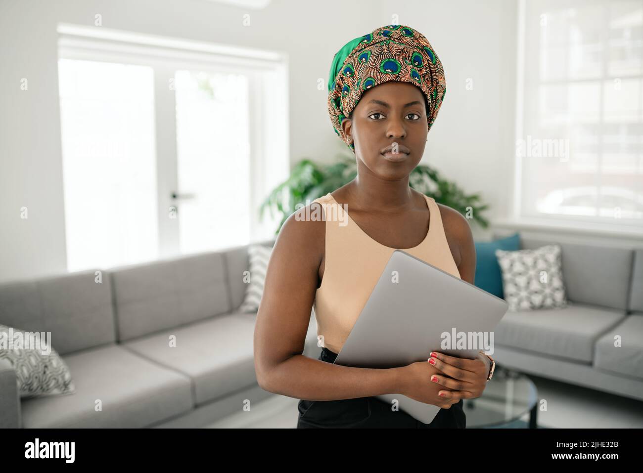 Portrait of beautiful smiling Black African woman standing in modern home with laptop in hand, wearing a traditional headscarf Stock Photo