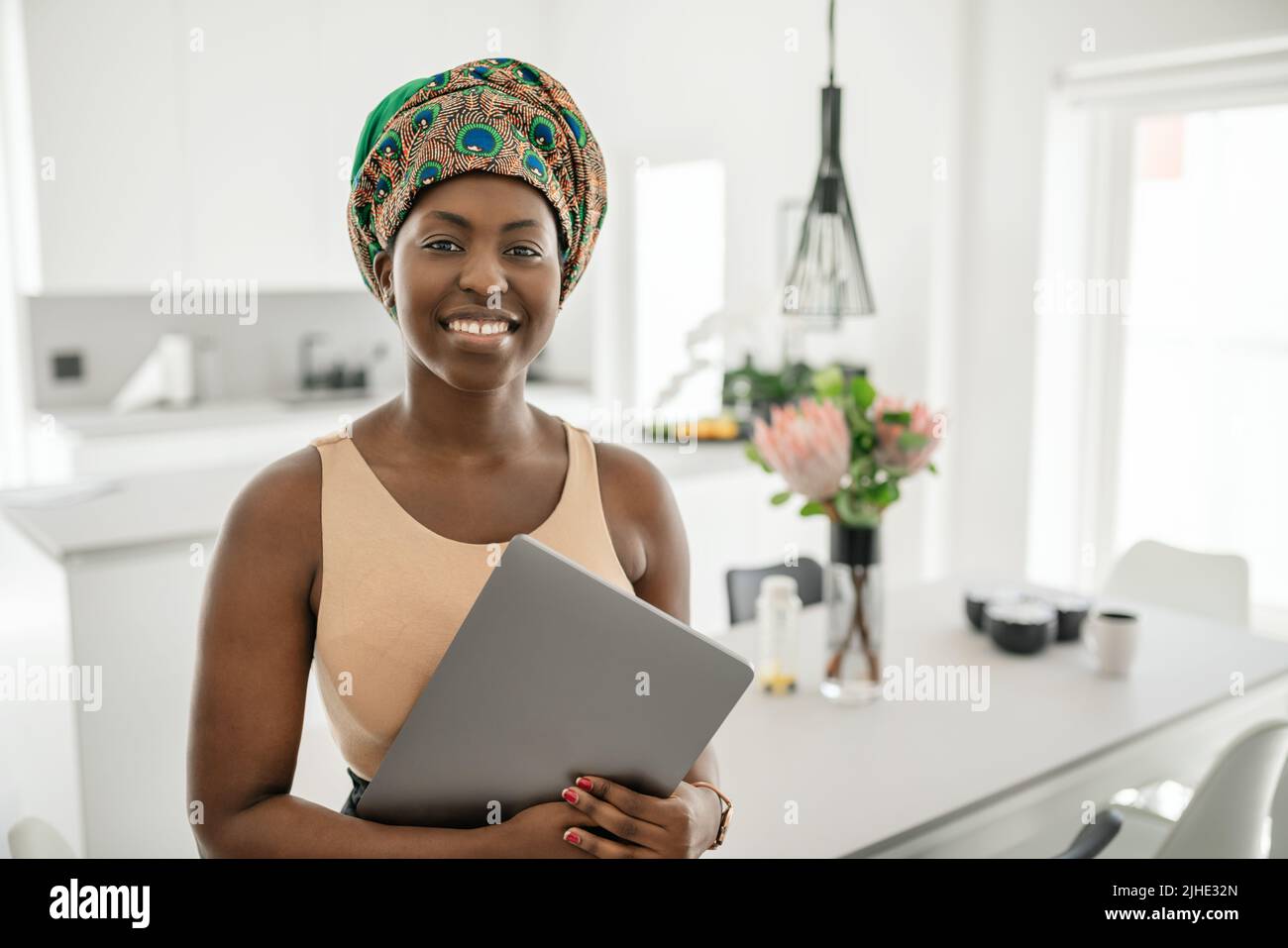 Portrait of beautiful smiling Black African woman standing in modern home with laptop in hand, wearing a traditional headscarf Stock Photo