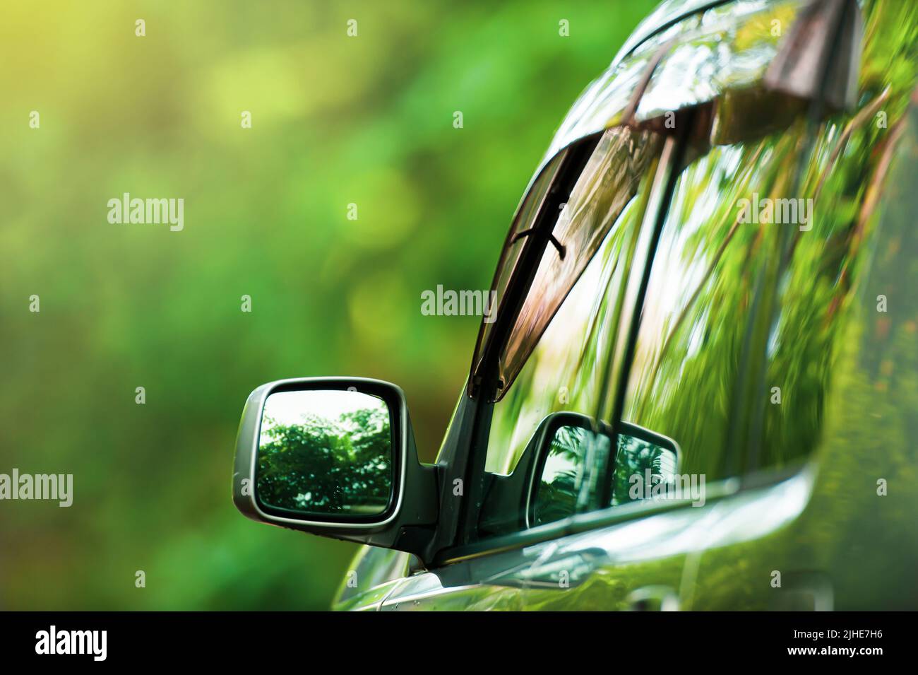 Rear view of a silver SUV car driving on a forest road, green trees reflected on the mirror, green forest blurred in the background. Road trip. Stock Photo