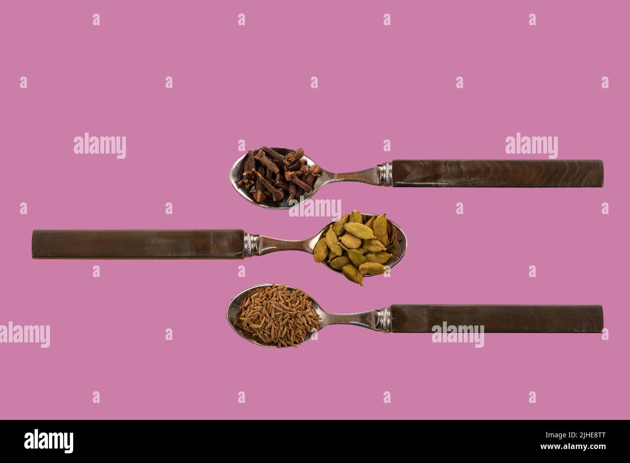 selection assorted indian spices cumin seeds cardamom cardamon pods cloves display on spoons on a colorful colourful pink lavender background Stock Photo