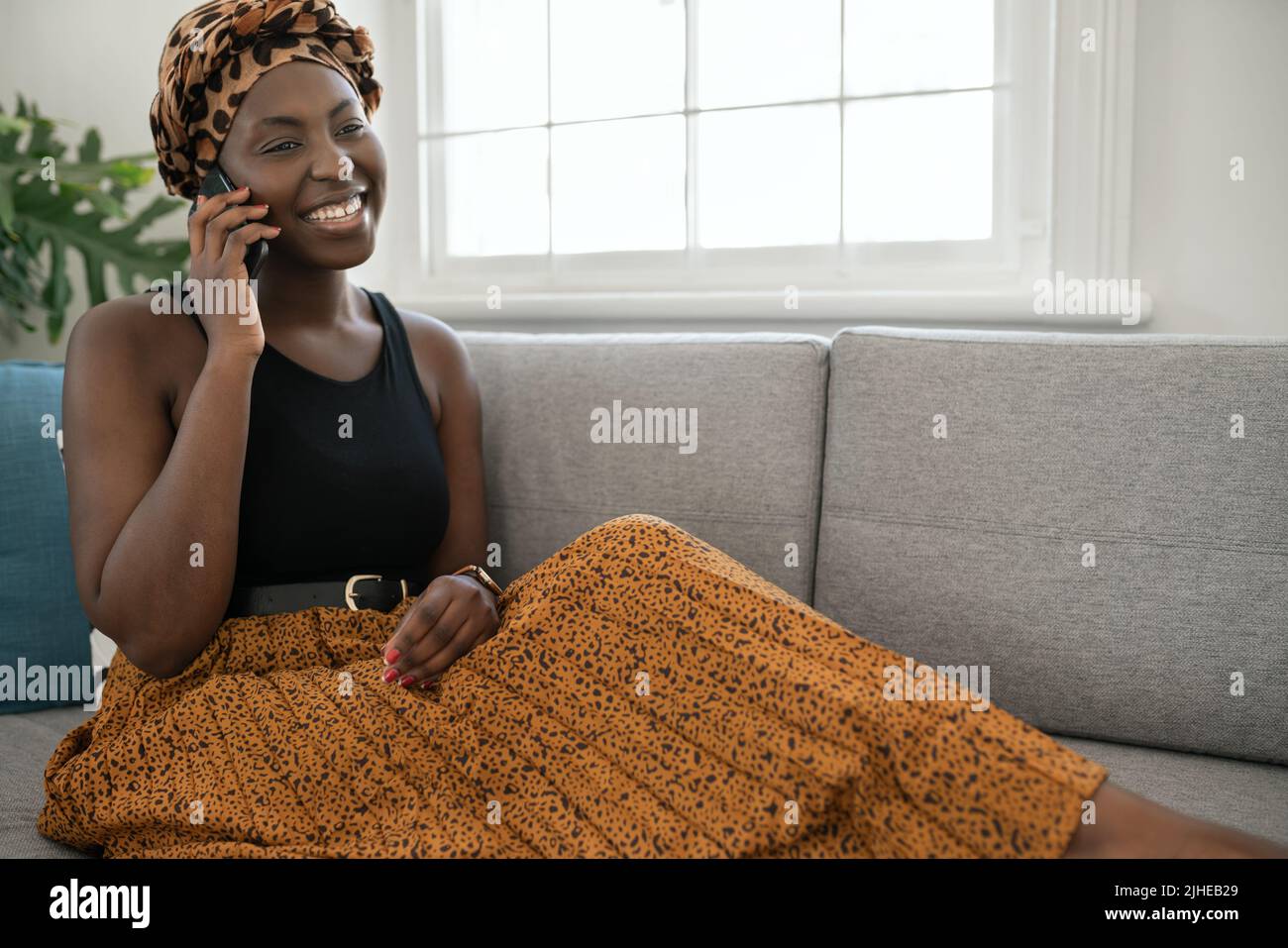 Beautiful traditional young Black gen z African woman sitting on sofa at home, talking smart phone, smiling, shot with copy space Stock Photo