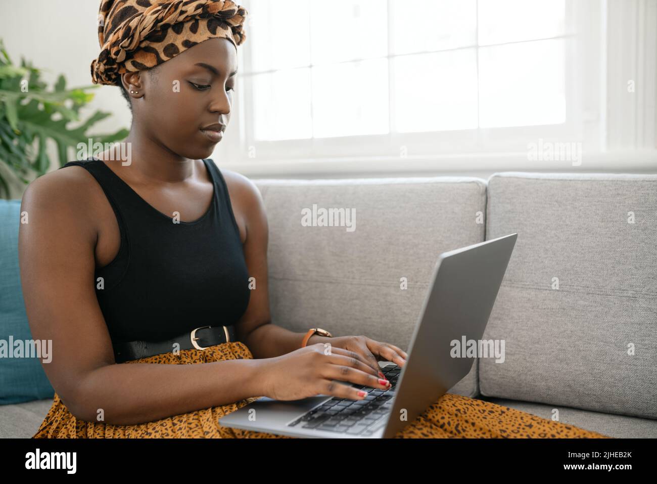 Close up Beautiful traditional young gen z African woman sitting on sofa at home, typing on laptop, smiling, shot with copy space Stock Photo