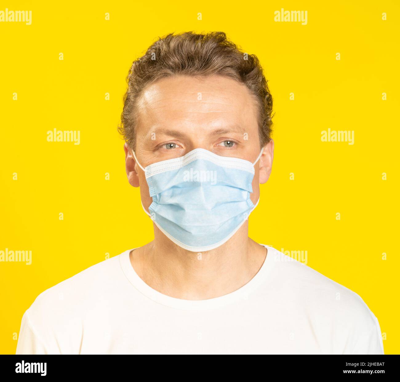 Sad face young man wearing a medical mask on his face. Sick young man in face mask wearing white t-shirt isolated on yellow background. A sick caucasian man in protective face mask.  Stock Photo