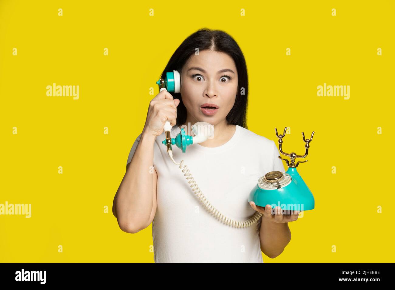 Shocked of winning lottery middle aged asian woman with vintage, retro telephone in hands excited, surprise face expression, isolated on yellow background. Communication concept.  Stock Photo
