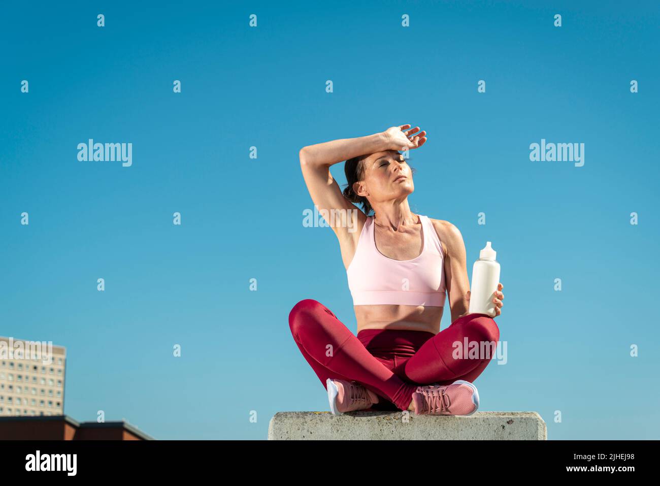 fit sporty woman sitting crossed legged in the sun holding a glass bottle of water, heatwave concept. Stock Photo