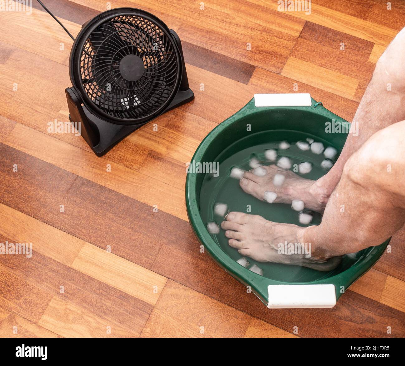 Man with feet in bowl of iced water next to fan during heatwave. Stock Photo