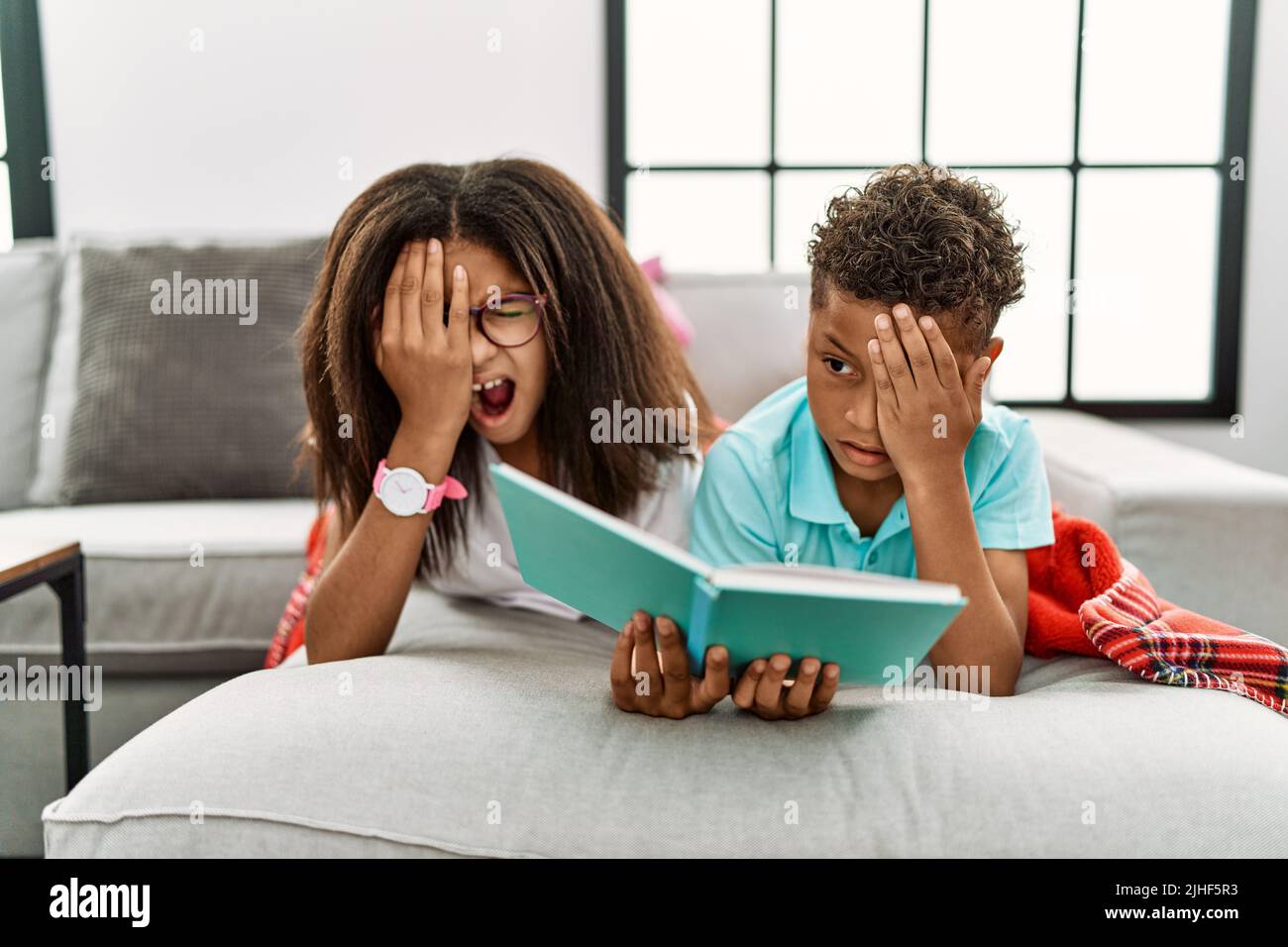 Two siblings lying on the sofa reading a book yawning tired covering