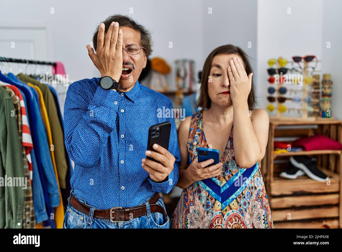 Middle age interracial couple at retail shop using smartphone yawning tired covering half face, eye and mouth with hand. face hurts in pain. Stock Photo