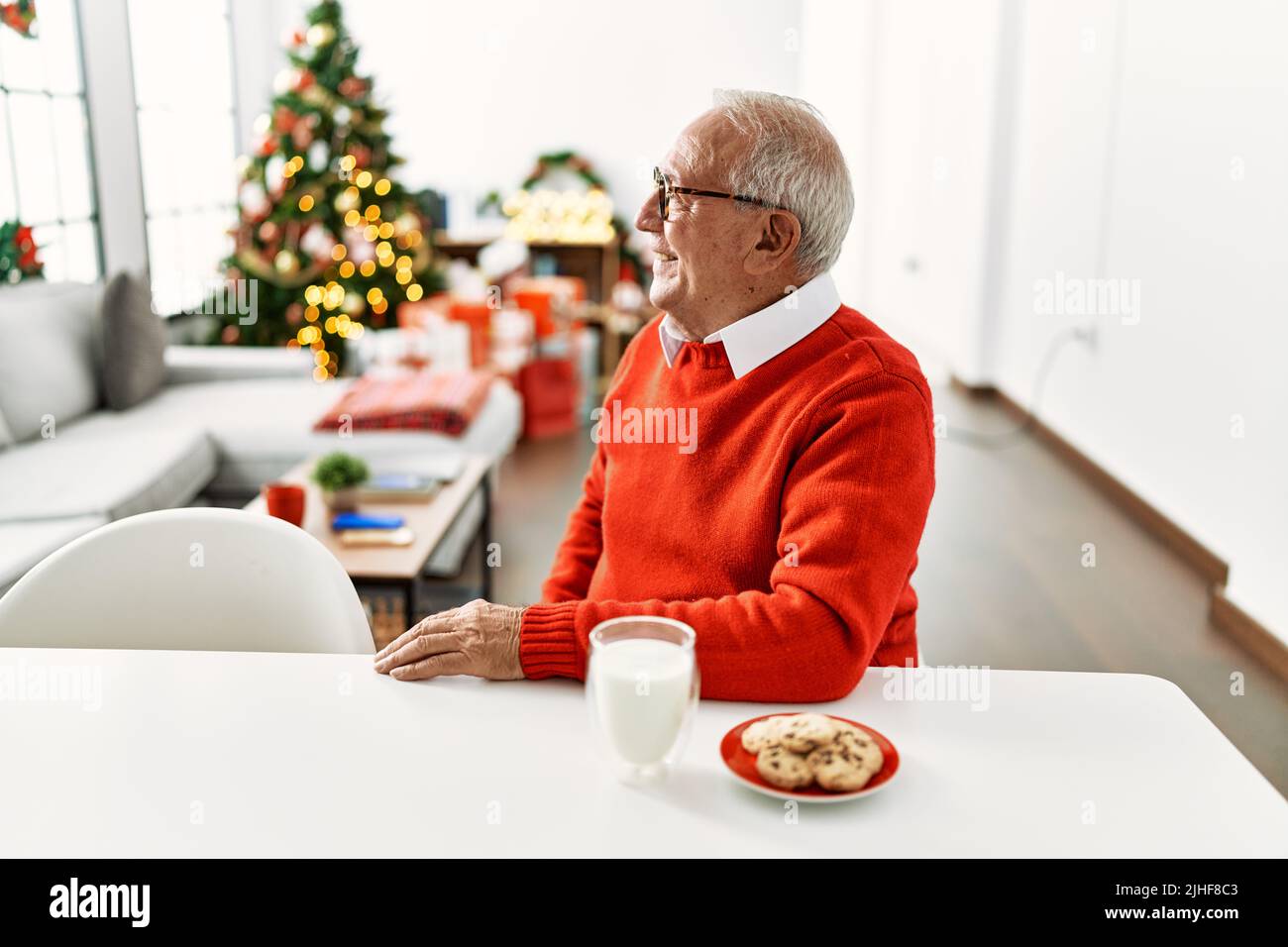 Senior man with grey hair sitting on the table with cookies by christmas tree looking to side, relax profile pose with natural face with confident smi Stock Photo