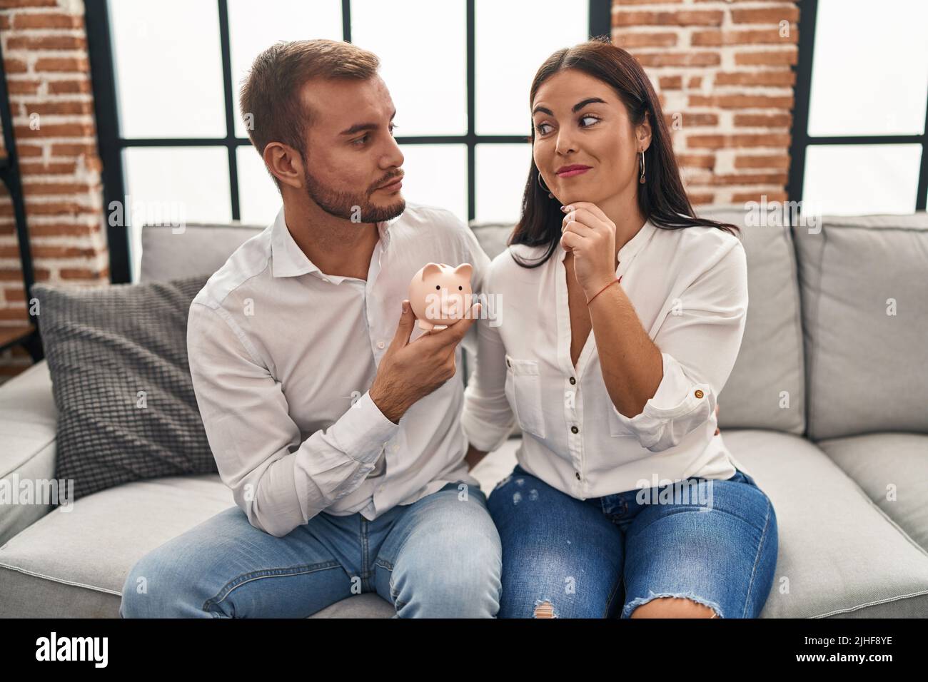 Young hispanic couple holding piggy bank sitting on the sofa serious ...