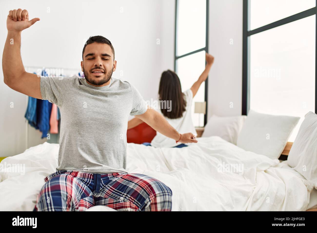 Young latin couple yawning and stretching arms sitting on the bed Stock