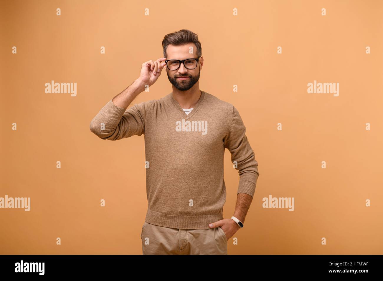 Charismatic bearded caucasian handsome man isolated on beige background standing and looks at the camera, male entrepreneur in casual wear adjusting stylish eyeglasses Stock Photo