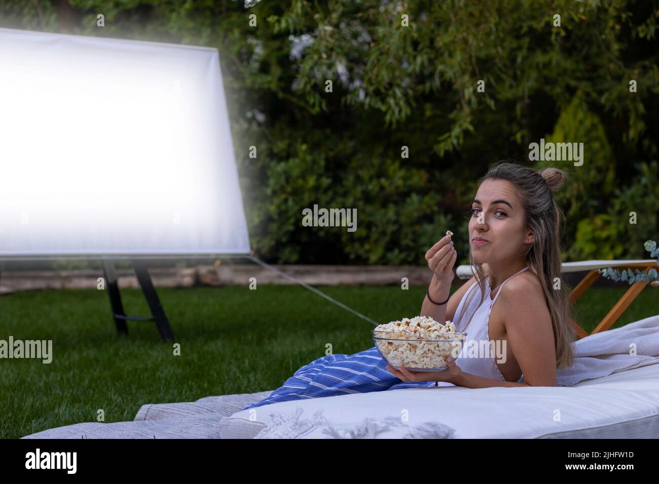 Young woman looking to camera watching a movie in the garden while showing the popcorn she's eating Stock Photo