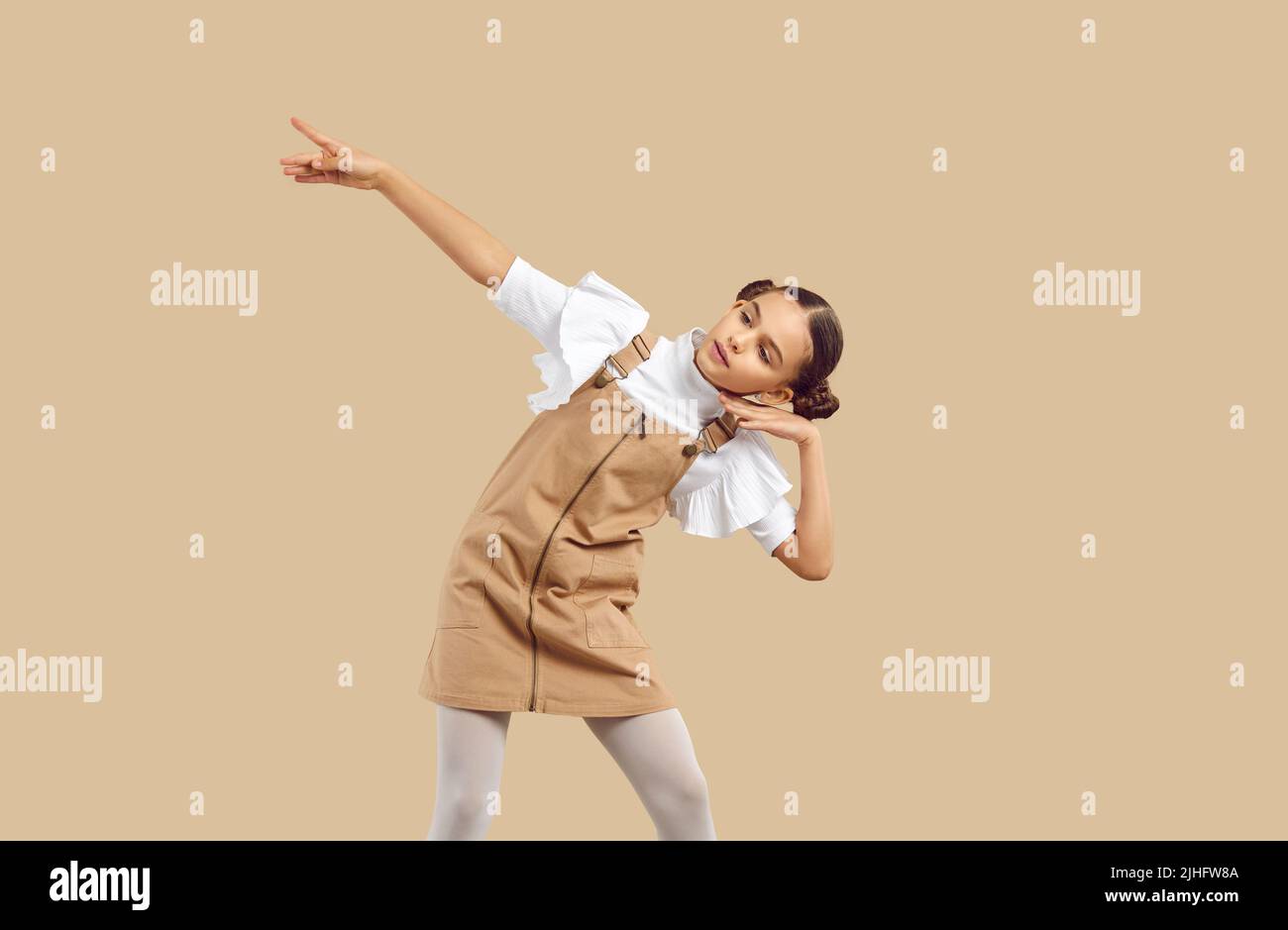 Little girl in casual beige denim pinafore dress dancing on beige studio background Stock Photo