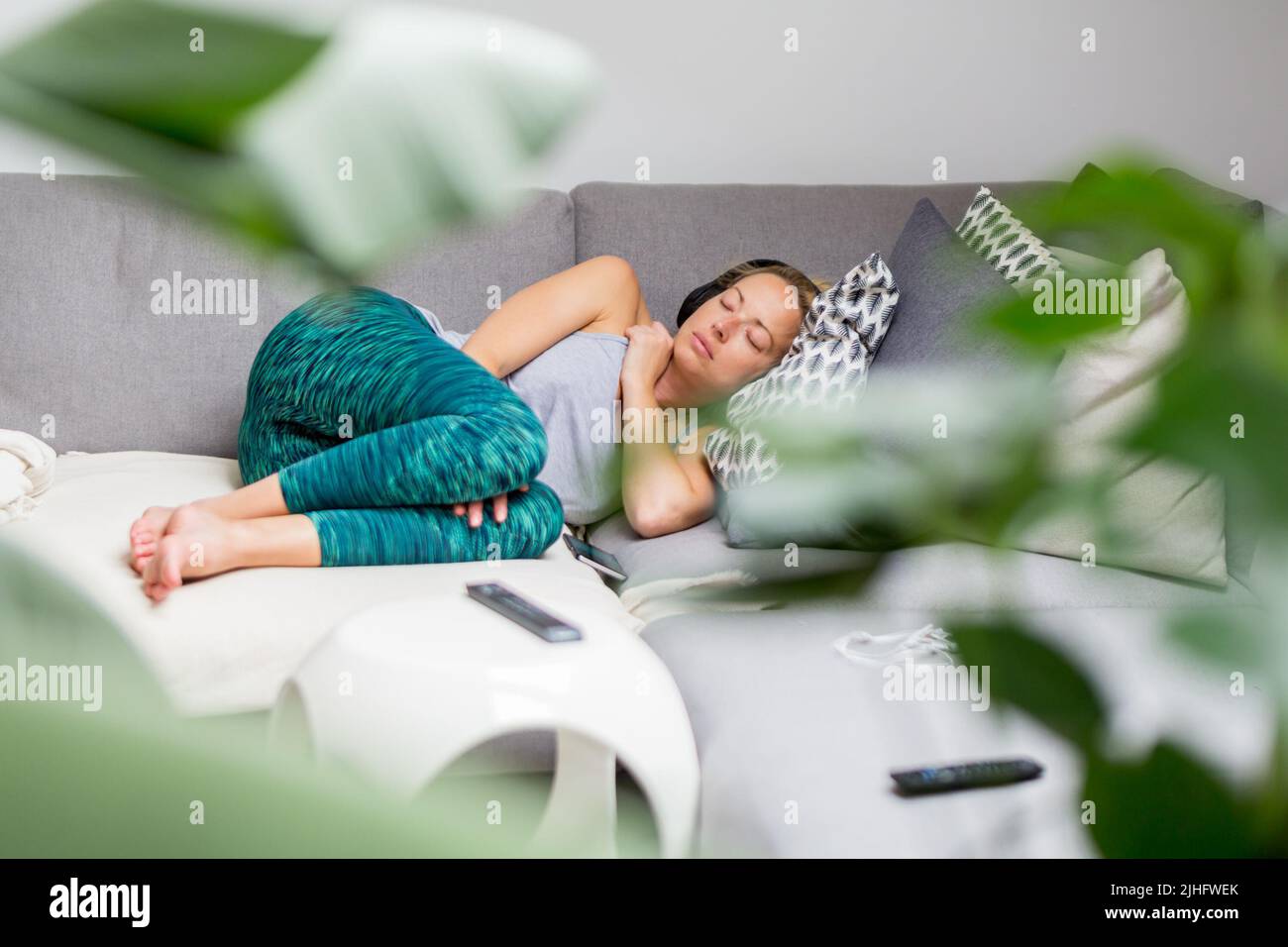 Young casual woman lying on couch cushion with eyes closed, relaxing on cozy sofa pillow, relaxed girl taking nap at home, wearing headphones Stock Photo