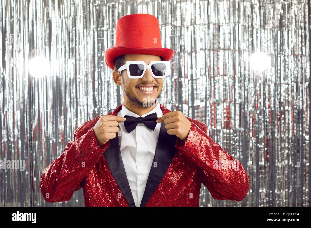 Funny guy in red sequin party suit, top hat and sunglasses smiles and fixes his bowtie Stock Photo
