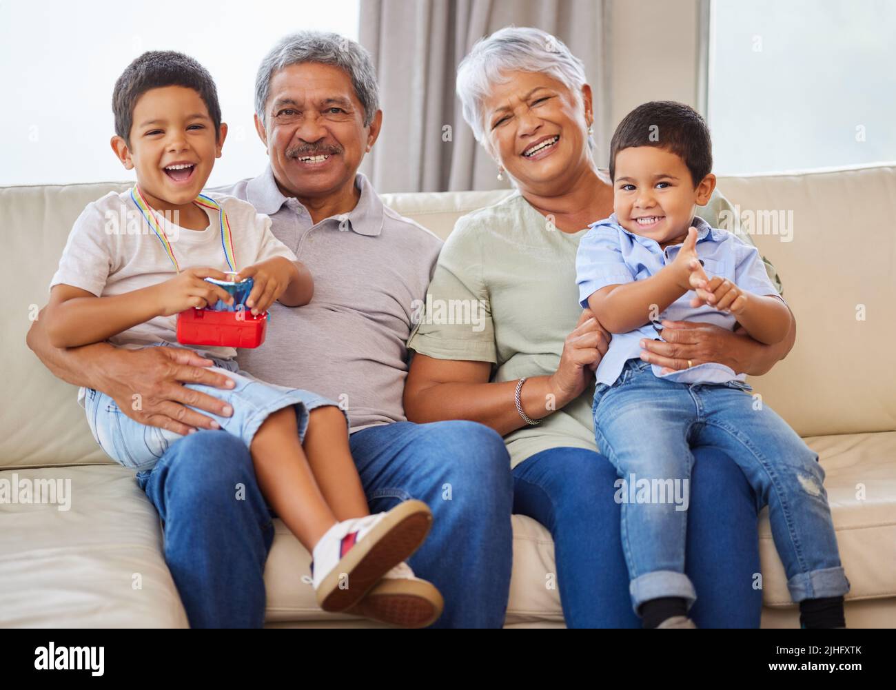 Portrait of smiling grandparents while holding their grandsons on a sofa in the lounge. Senior hispanic man and woman spending time with their Stock Photo