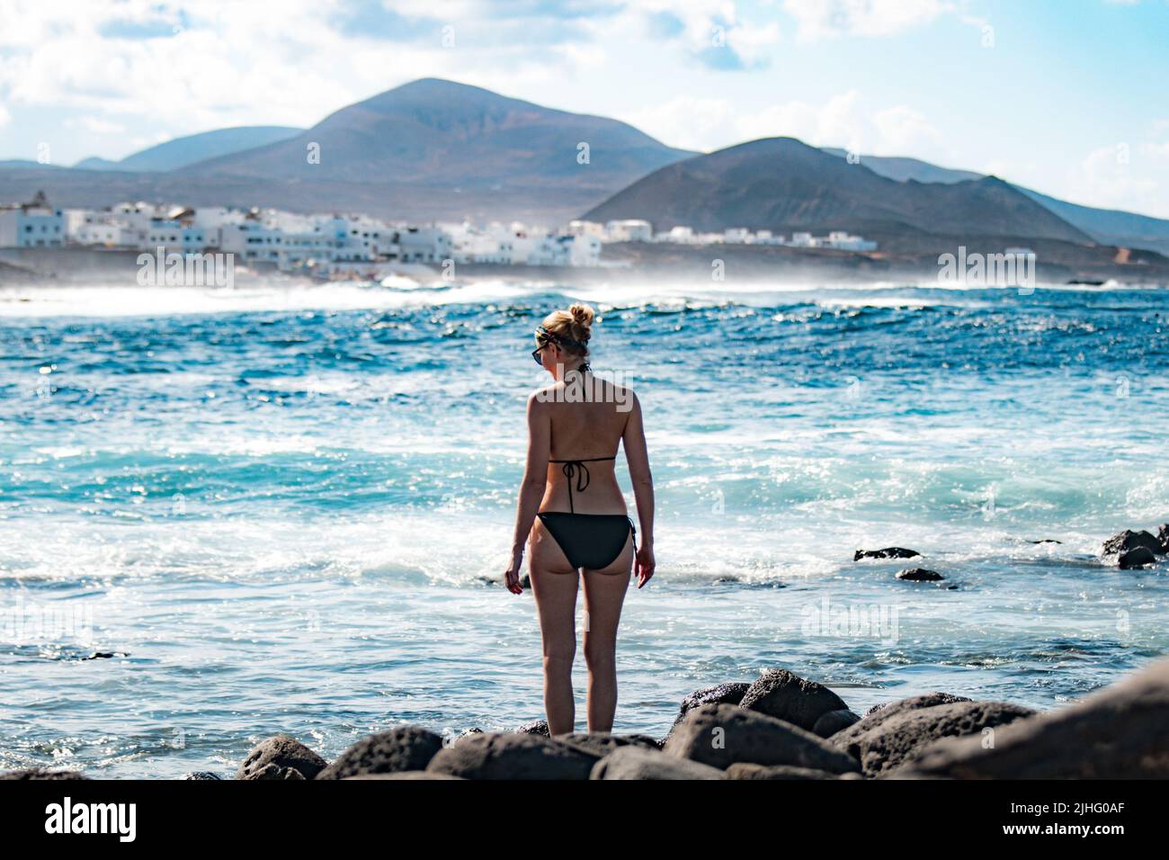 Female tourist at wild rocky beach and coastline of surf spot La Santa Lanzarote, Canary Islands, Spain. La Santa village and volcano mountain in Stock Photo