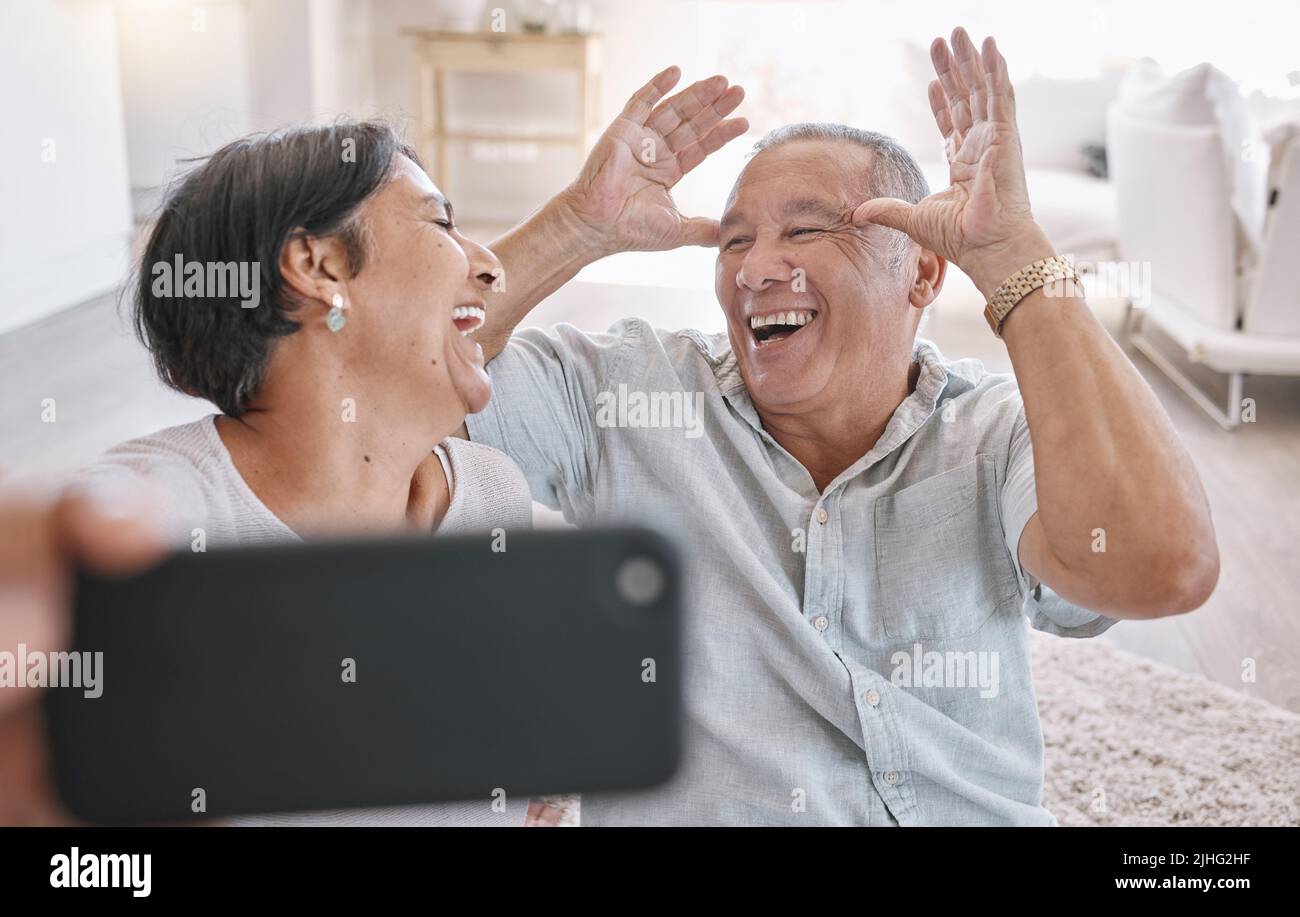 Closeup of an affectionate senior couple taking selfies while relaxing in the living room at home. Mixed race couple having fun taking photos in the Stock Photo