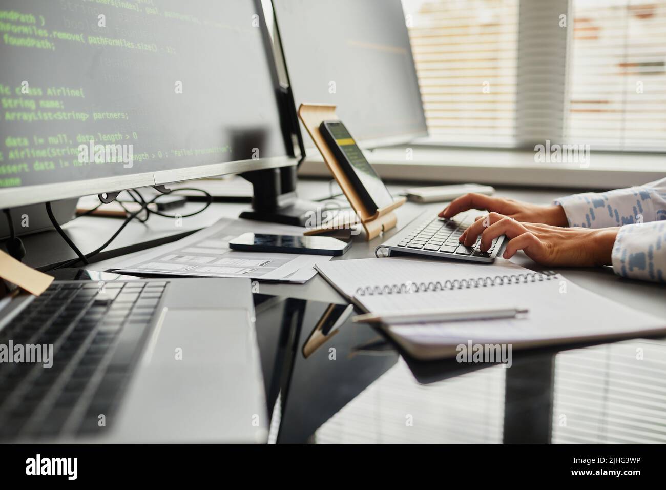 Close-up of female programmer typing code on keyboard from smartphone while sitting at table in front of computer monitors at office Stock Photo