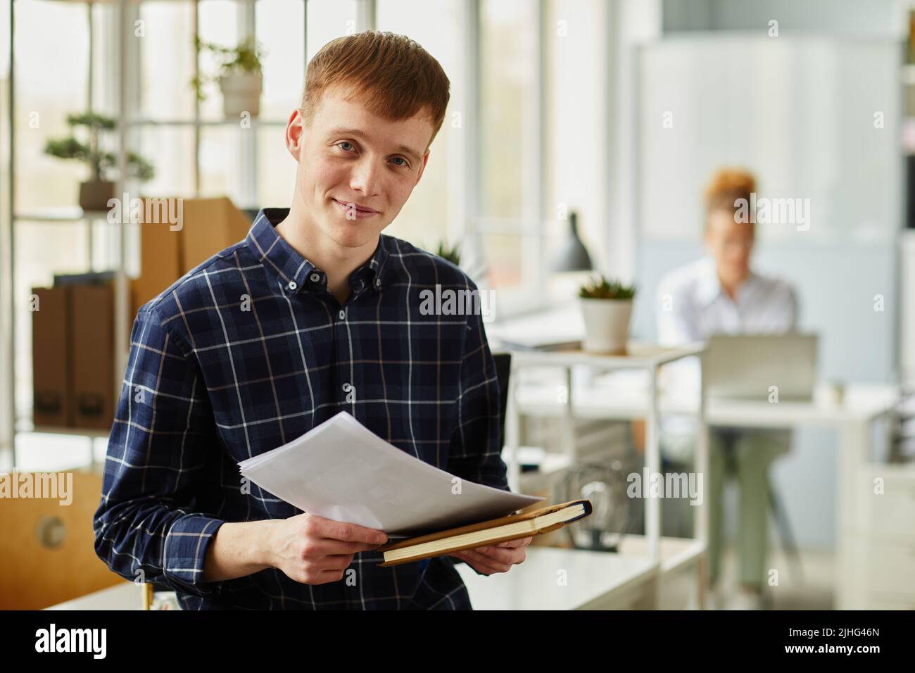 Portrait of young programmer in checked shirt holding notebook and documents and looking at camera while standing at office Stock Photo