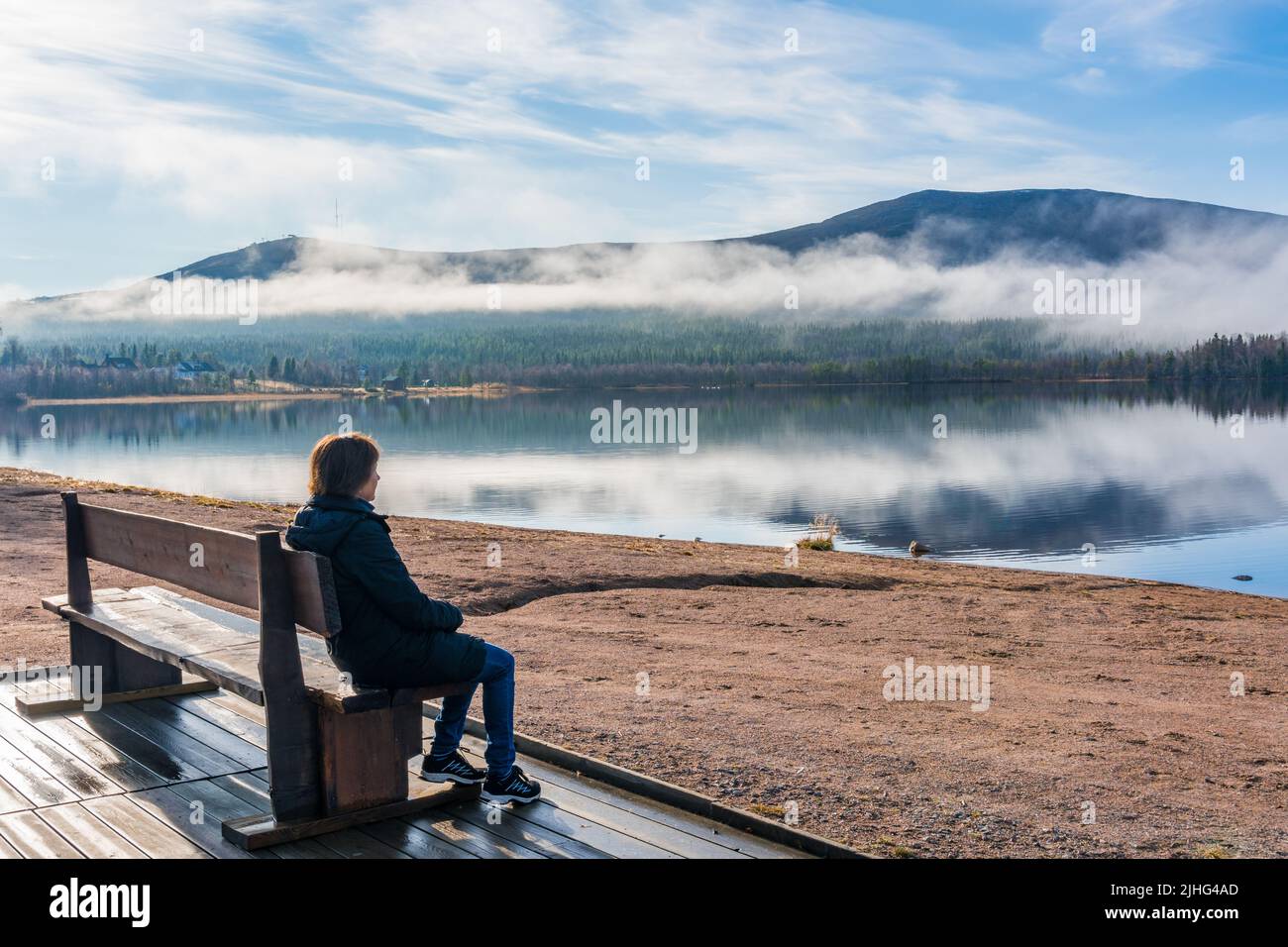 Woman sitting on a bench at a beach watching the nice view with mountain and river, Gällivare, Swedish Lapland, Sweden Stock Photo