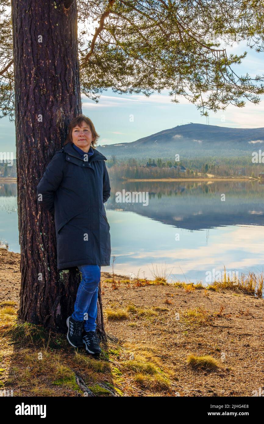 Woman standing on a beach by a pine tree in autumn season with mountain and fog in background, Gällivare, Swedish Lapland, Sweden Stock Photo