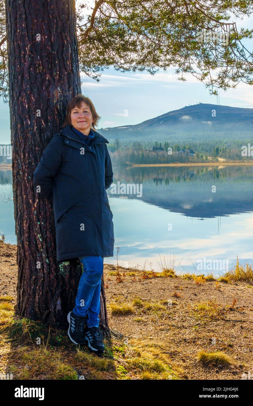 Woman standing on a beach by a pine tree in autumn season with mountain and fog in background, Gällivare, Swedish Lapland, Sweden Stock Photo