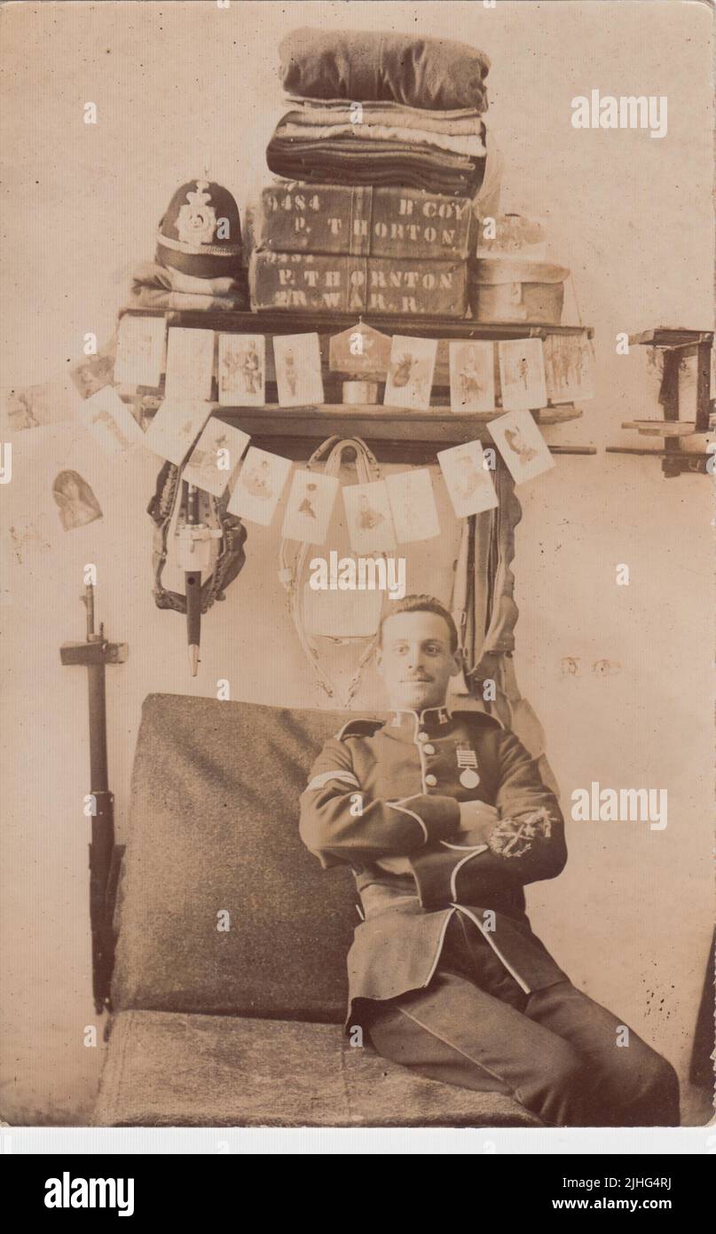 Philip Thornton, Royal Warwickshire Regiment, photographed in his barracks bed space in Portland. Thornton is in uniform, wearing a medal, and has his army helmet, towels or bed linen, and cases on shelves behind him. His rifle is on the wall to the left of the bed and postcards and photos (most with a military theme) are hung from the shelves. This photographic postcard was sent to his future wife Hilda White in 1905. Thornton was killed in France on 13 October 1914, aged 33, during the first months of the First World War Stock Photo