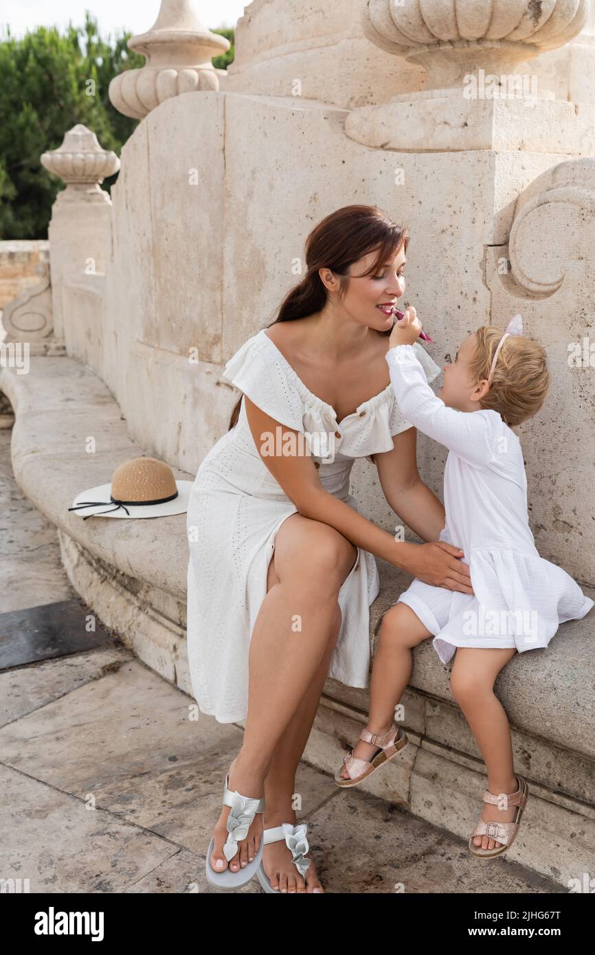 Toddler girl applying lip gloss on mother lips on stone bench of Puente Del Mar bridge in Valencia Stock Photo