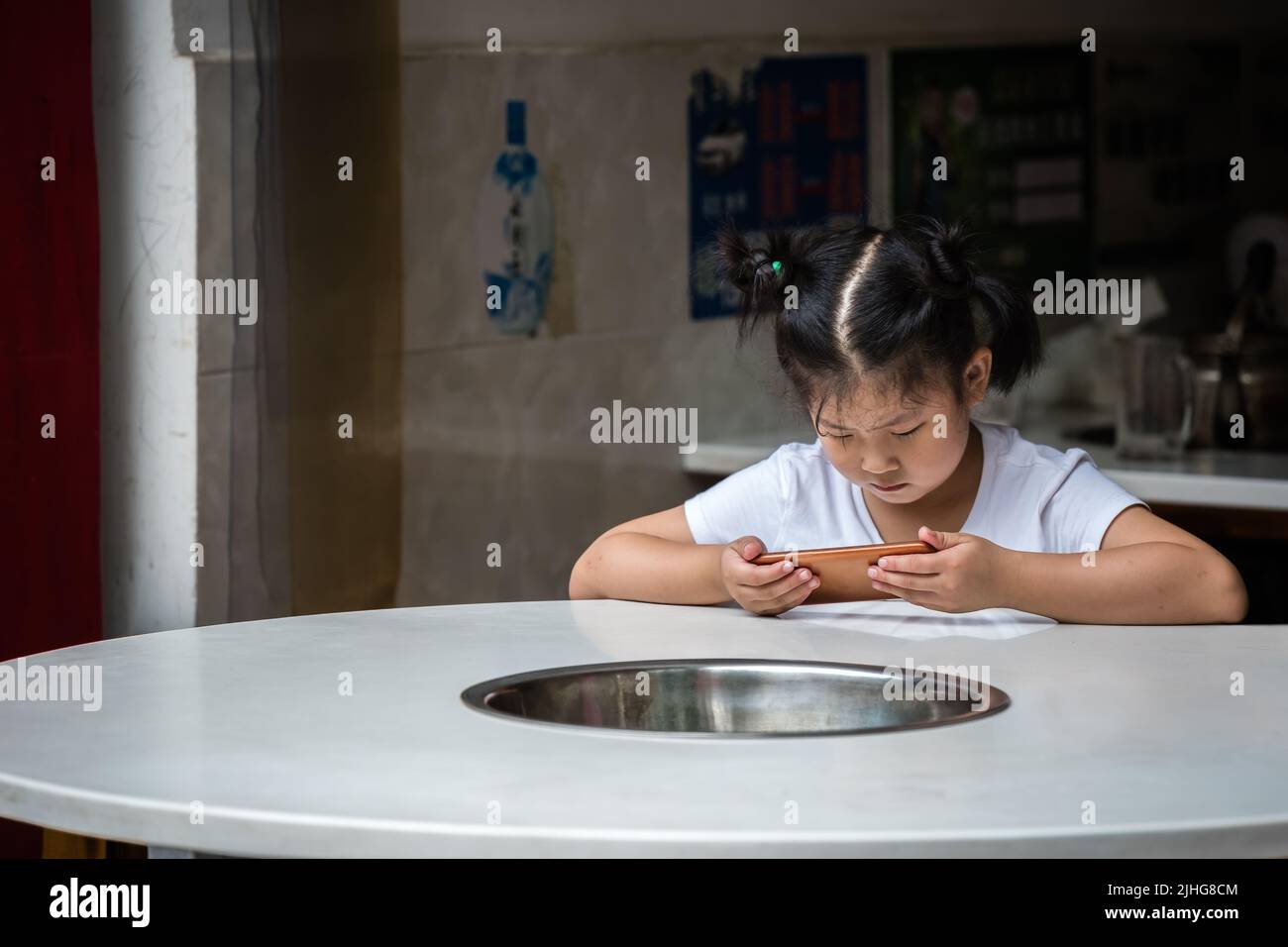 Zigong, China - July 2019 : Young little chinese girl sitting by a round table writing message and texting on her mobile phone Stock Photo