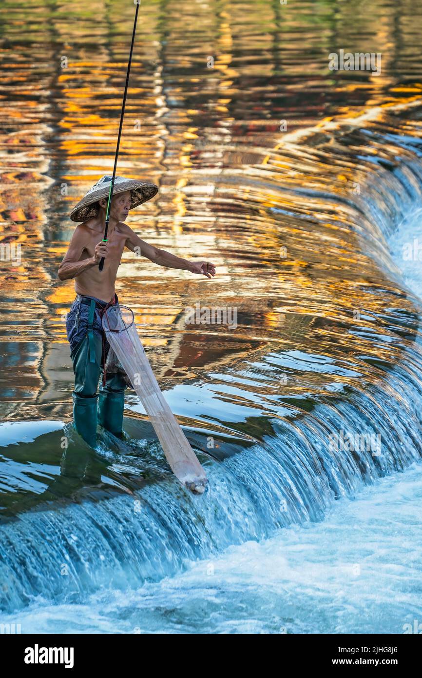 Feng Huang, China -  August 2019 : Fisherman with a traditional triangular chinese hat standing in waters of Tuo river flowing through the centre of F Stock Photo