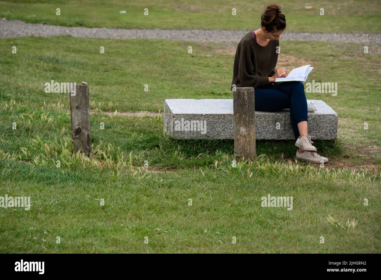 London, UK - June 2020 : Young woman sitting on a concrete slab bench and reading a book on a hot summer day Stock Photo