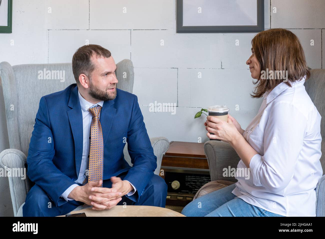 A young woman in a white shirt and jeans holding a mug of coffee and a middle-aged Arab man in a business suit talking at a cafe table. Coworking. Stock Photo