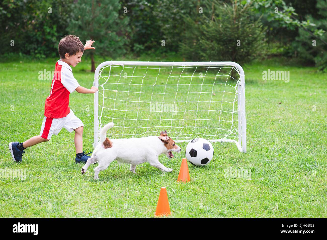 Kid boy in red and white kit playing football (soccer) with his dog at training ground Stock Photo