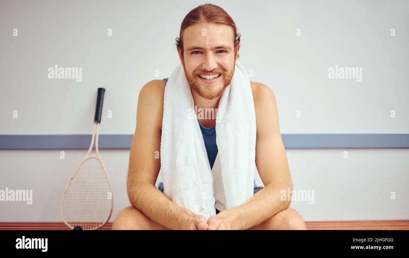 Portrait of a squash player with his racket. Caucasian man sitting in a gym locker room relaxing. Fit, young athlete taking a break before his match Stock Photo